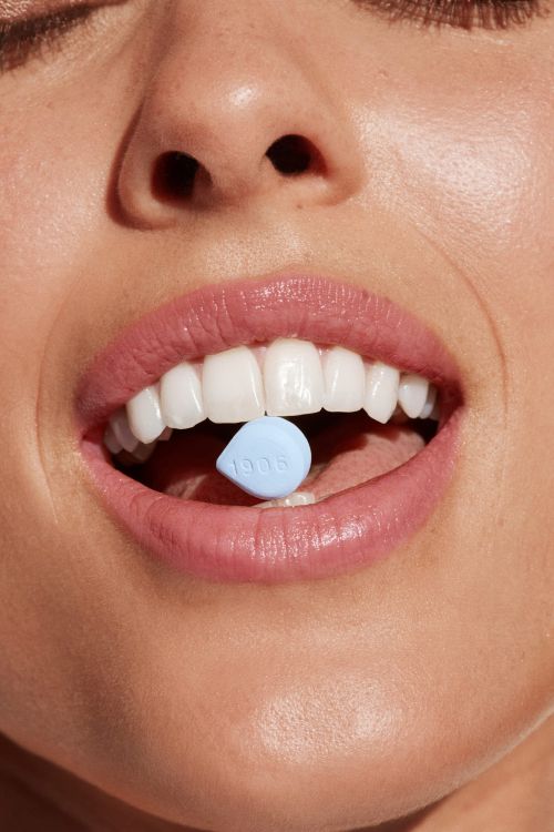 Close-up of a woman's face with pink lipstick and white teeth, holding a small blue pill with the number '1906' on her tongue.