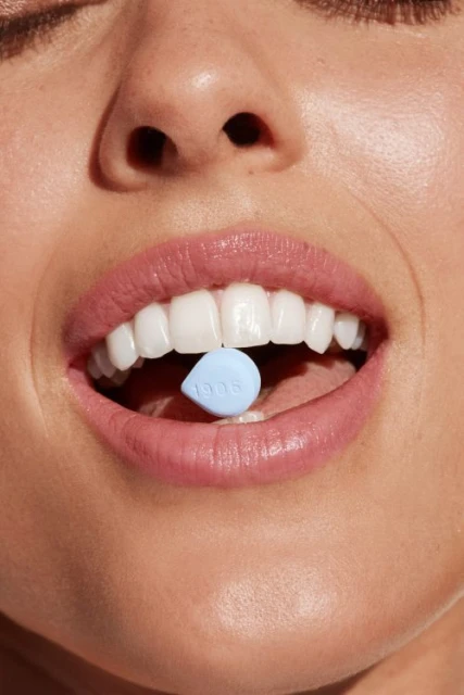 Close-up of a woman's face with pink lipstick and white teeth, holding a small blue pill with the number '1906' on her tongue.