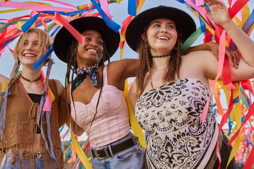 Three young women smiling and enjoying themselves surrounded by colorful streamers, dressed in casual fashion with hats and summer tops.