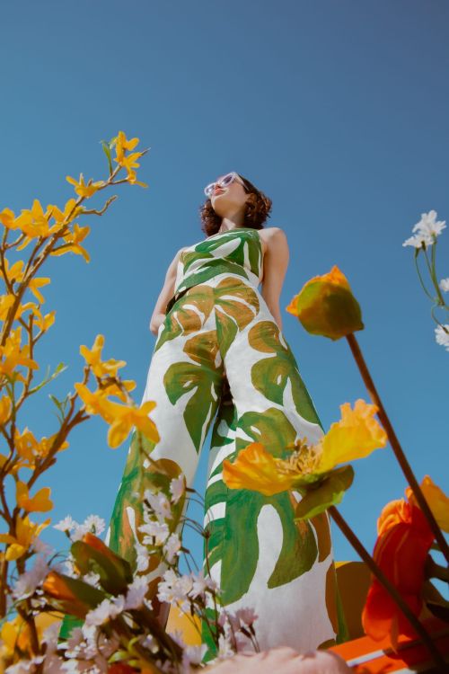 Low angle shot of a woman wearing a green and white leaf print outfit against a vibrant blue sky with foreground flowers.