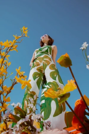 Low angle shot of a woman wearing a green and white leaf print outfit against a vibrant blue sky with foreground flowers.