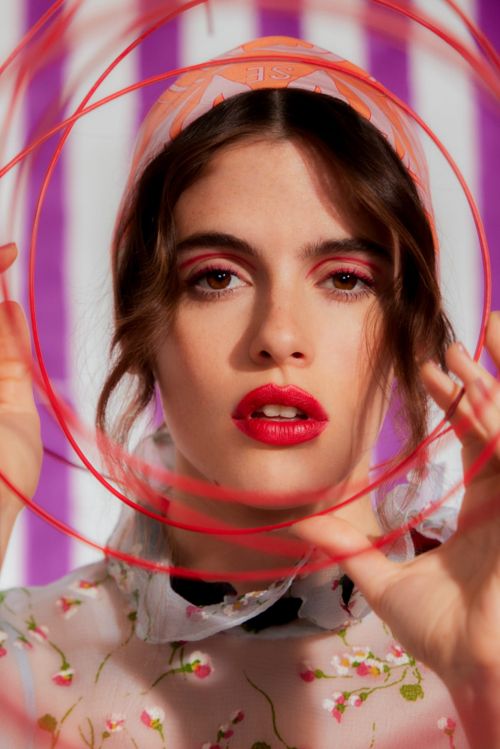 Close-up portrait of a young adult woman with red eye makeup and lipstick, wearing a floral top and headscarf, holding red wire circles.