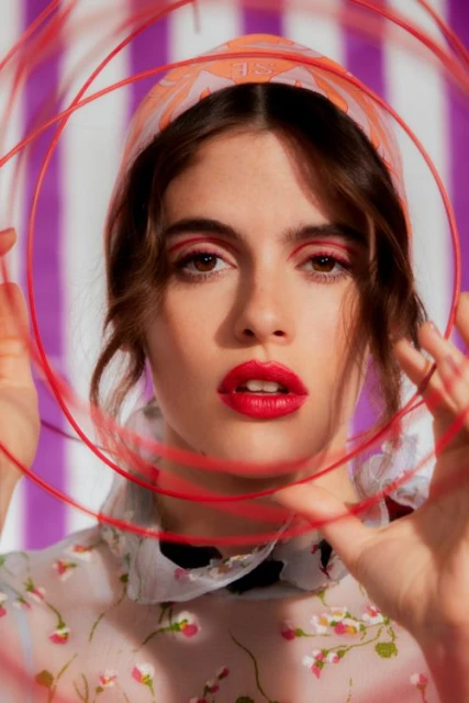 Close-up portrait of a young adult woman with red eye makeup and lipstick, wearing a floral top and headscarf, holding red wire circles.