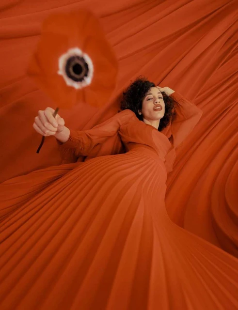 Woman lies on a red, draped background, holding an orange flower. She wears a red dress with long sleeves and has curly, dark hair.