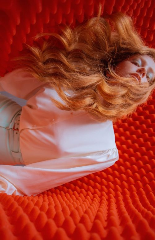 A woman with long, auburn, wavy hair and a light-colored top reclines on a textured, orange surface in a close-up, eye-level studio shot.