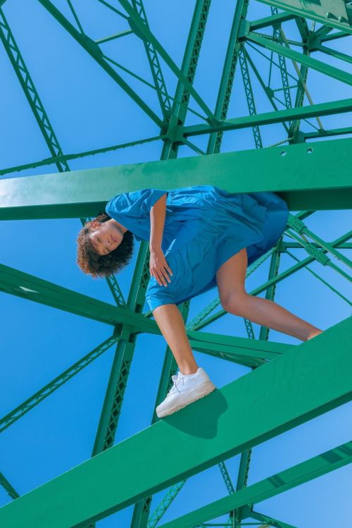 A woman in a blue dress stands on a green steel bridge against a blue sky, with white sneakers and a striking pose.