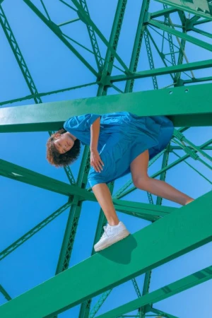 A woman in a blue dress stands on a green steel bridge against a blue sky, with white sneakers and a striking pose.