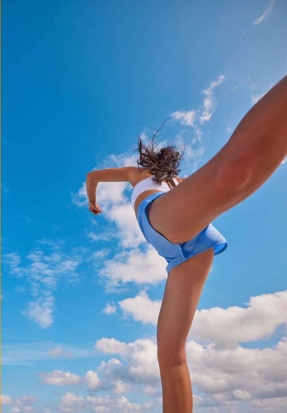 A low-angle shot captures a woman with dark hair outdoors, wearing a white sports bra and blue shorts, against a bright blue, cloudy sky.
