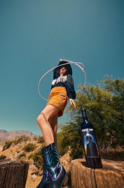 Woman wearing cowboy boots and hat poses near a bottle of wine with desert mountains and blue sky in background for fashion and beverage.