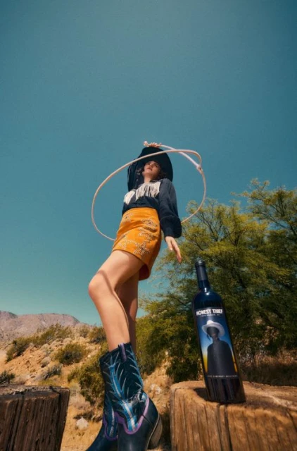 Woman wearing cowboy boots and hat poses near a bottle of wine with desert mountains and blue sky in background for fashion and beverage.