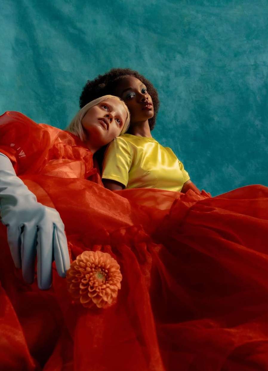 Two women, one with blonde hair and one with afro hair, in bright orange and yellow dresses, and a large orange flower. 