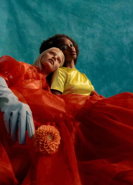Two women, one with blonde hair and one with afro hair, in bright orange and yellow dresses, and a large orange flower. 