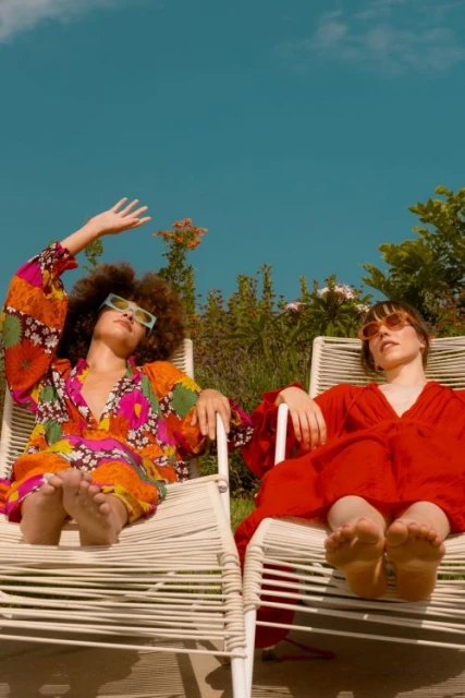 Two women in colorful dresses lounge on deck chairs outdoors under a clear sky surrounded by green plants, enjoying a sunny day.