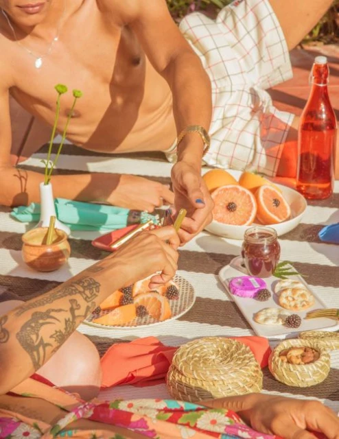 Two people enjoying a sunny picnic with sliced grapefruits, pastries, bottled beverage and small flower vases on a striped blanket.