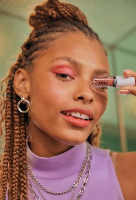 Beautiful young adult female with braided hair smiles while holding a cosmetic product. She is wearing pink eyeshadow and silver jewelry.