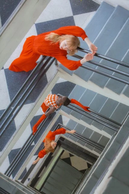Fashion shot from above features two women on a staircase, one in red dress, one in checkered top and orange leggings, facing up.