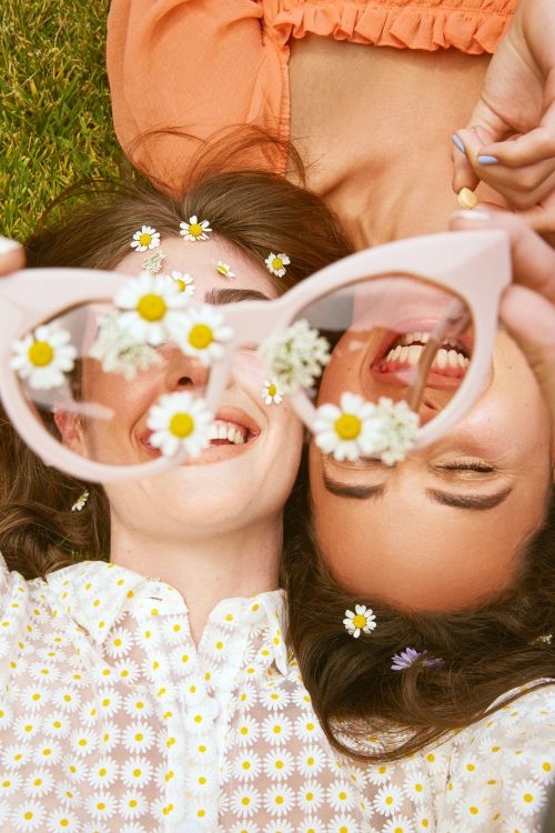 Two smiling women lie head-to-head on green grass, one holding sunglasses adorned with daisies in an eye-level, medium outdoor shot.