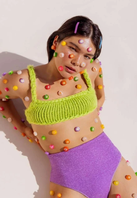 Brunette woman with colorful adornments posing against a white wall in a bright knitted crop top and sparkling lilac high-waisted shorts.