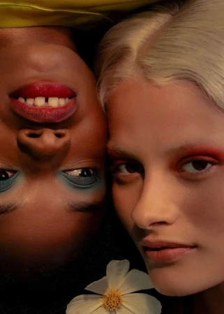Close-up portrait of two women lying head to head with vibrant makeup and a white flower nearby photographed by Jimmy Marble.