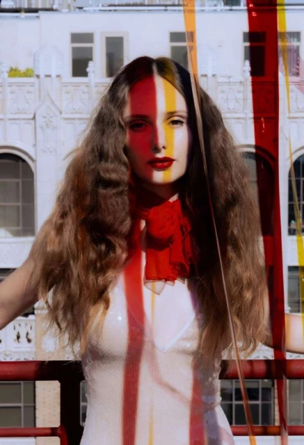 A woman with long brown hair stands in front of a white building, wearing a white dress and red scarf, color stripes overlaying her face.
