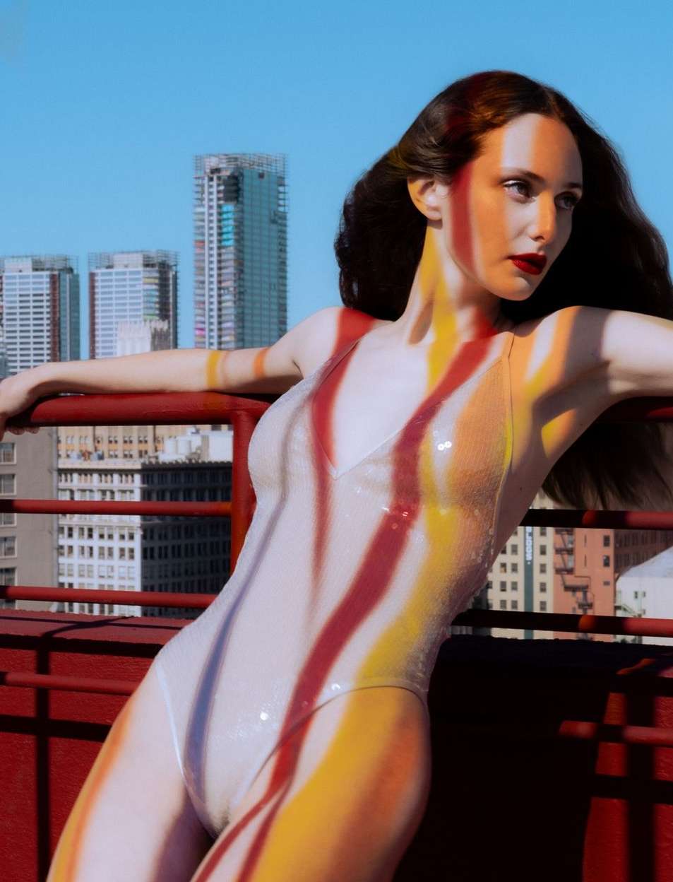 A woman in a swimsuit poses on a rooftop with a city skyline behind her. Her body has red and yellow paint stripes.