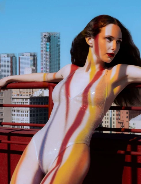 A woman in a swimsuit poses on a rooftop with a city skyline behind her. Her body has red and yellow paint stripes.