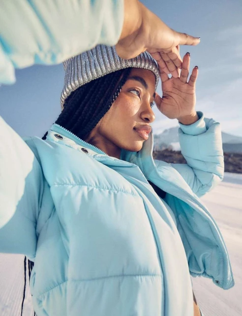 A young adult woman in a light blue puffer jacket and silver beanie gazes into the distance on a snowy, sunny day.