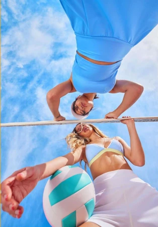 Two women in swimwear pose with a volleyball under a blue, partly cloudy sky, shot from a low angle capturing a summery, sporty aesthetic.