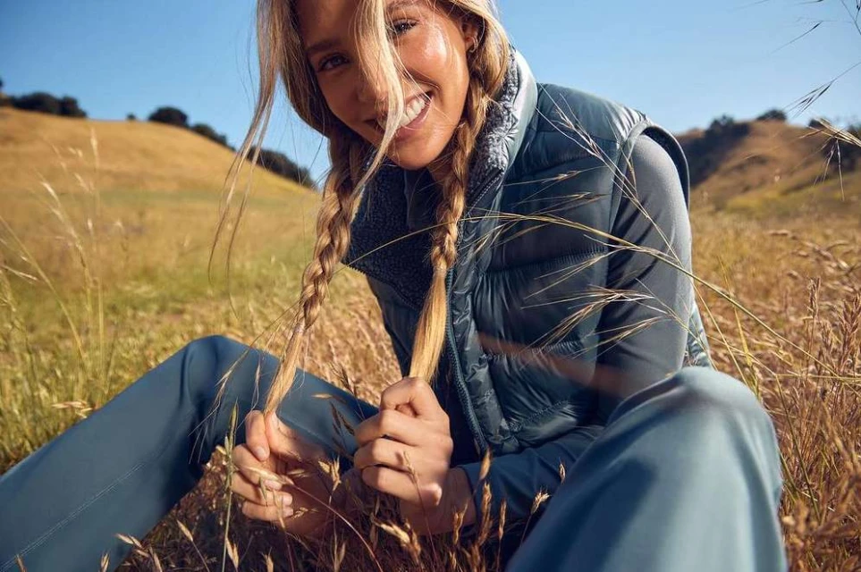 Woman with blonde braids in a gray vest sits in a grassy field under a clear sky, smiling while holding her braids.