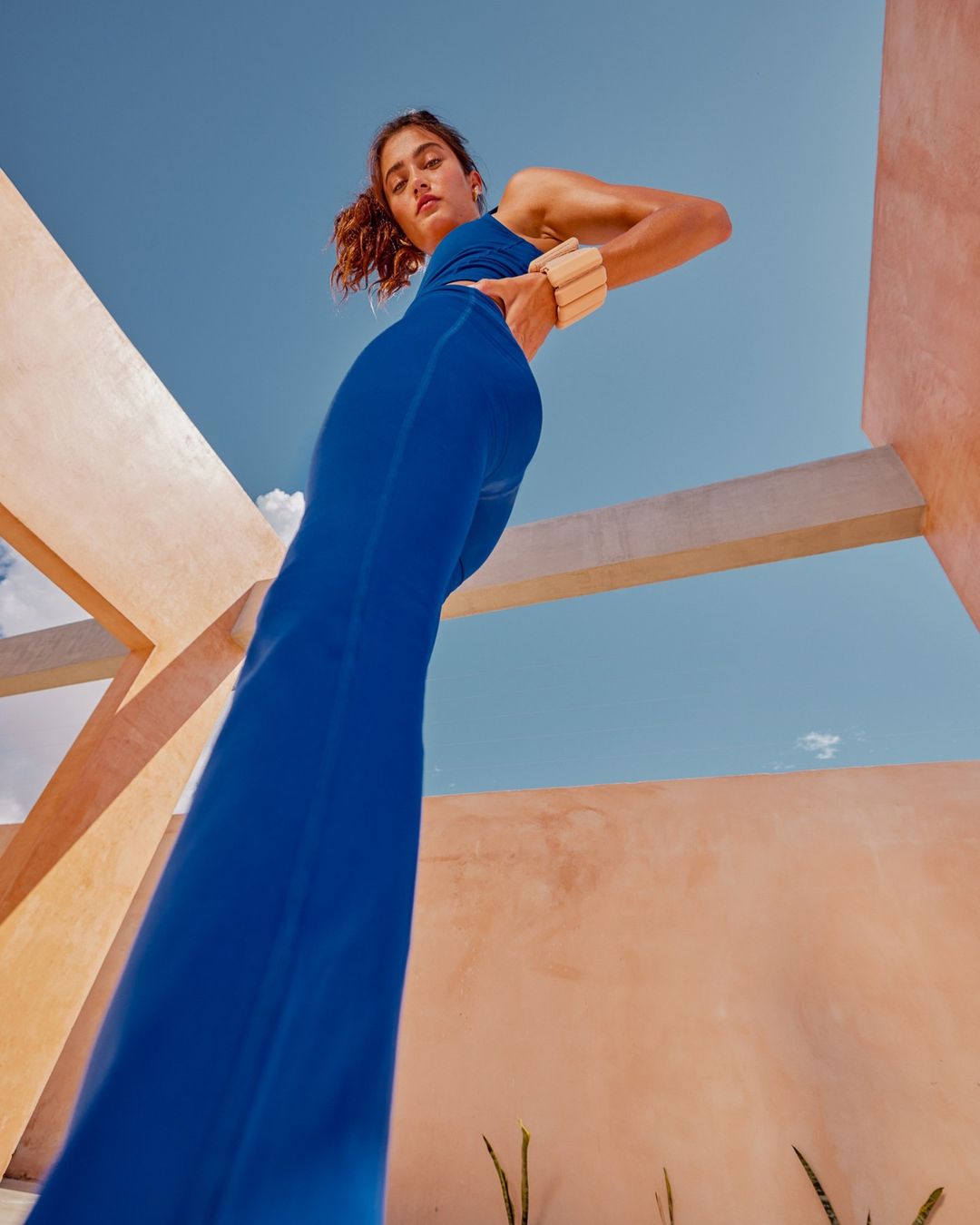 Low angle of a female model in a blue bodysuit posing near a peach-colored building against a clear sky, showcasing fitness and fashion.