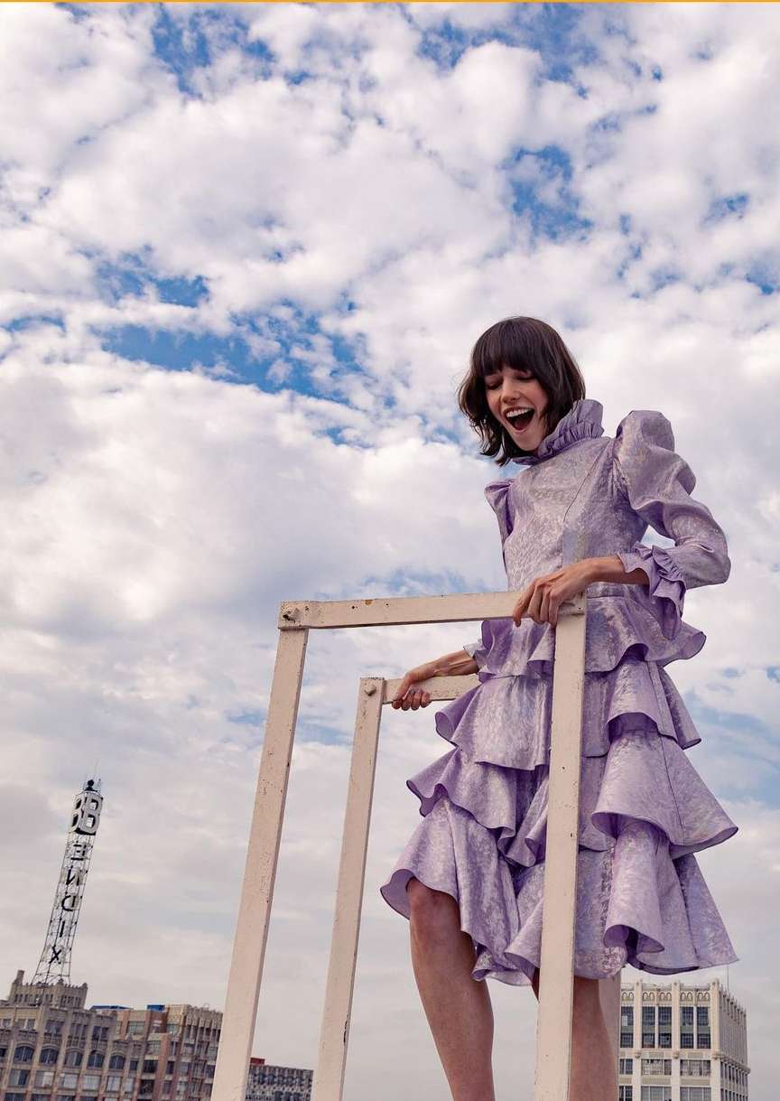 A light-skinned woman in a tiered lavender dress smiles on a rooftop, supported by a white frame against a cloudy sky and cityscape.