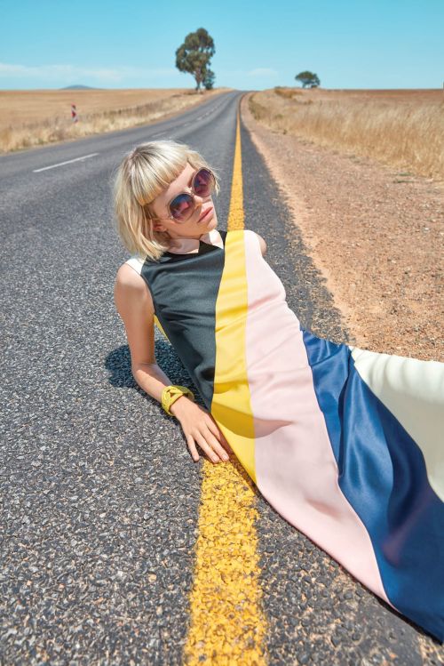 A blonde woman in sunglasses and a color-block dress lies on an asphalt road on a sunny day in a rural landscape, fashion editorial.