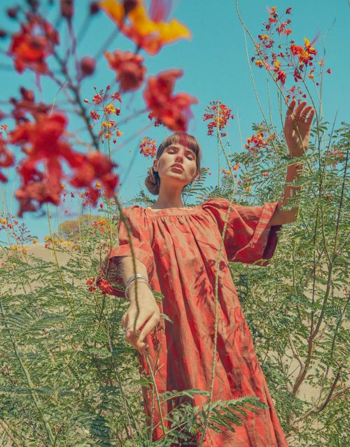 Woman in orange dress stands among orange and yellow flowers under blue sky, posing with raised hands in an outdoor setting.