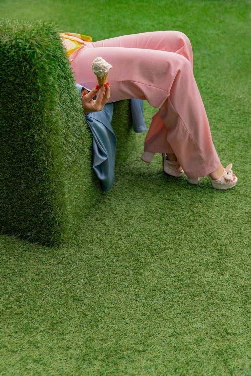 A person in pink pants sits on a chair covered in green grass holding an ice cream cone in an outdoor, grassy setting.