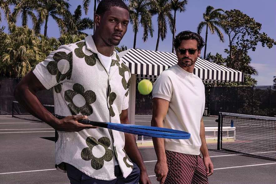 Two men pose on a tennis court with palm trees; one holds a racket and a tennis ball is in the air. Fashion editorial shot.