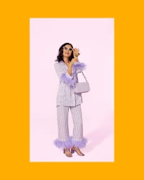 Woman in plaid pantsuit with feather cuffs holding glasses and a handbag against a pink background framed by yellow borders.