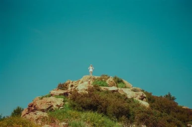 A small figure standing atop a rocky hill under a vast teal sky captured in a wide landscape shot by Jimmy Marble.
