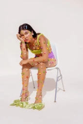 Woman seated on a white chair decorated with colourful candy pieces covering her skin and clothing against a plain background