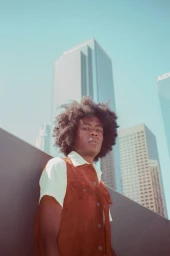 Man with afro wearing a brown vest and white shirt outdoors against a city skyscraper background by Jimmy Marble