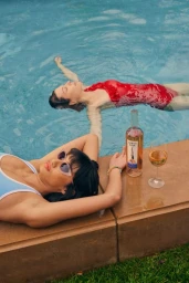 Two women relaxing by a swimming pool with a bottle of wine and a glass on the pool edge, photographed by Jimmy Marble.