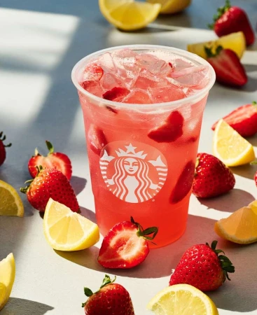 Strawberry lemonade beverage in a clear plastic cup with fresh strawberries and lemon slices scattered on a light grey surface.