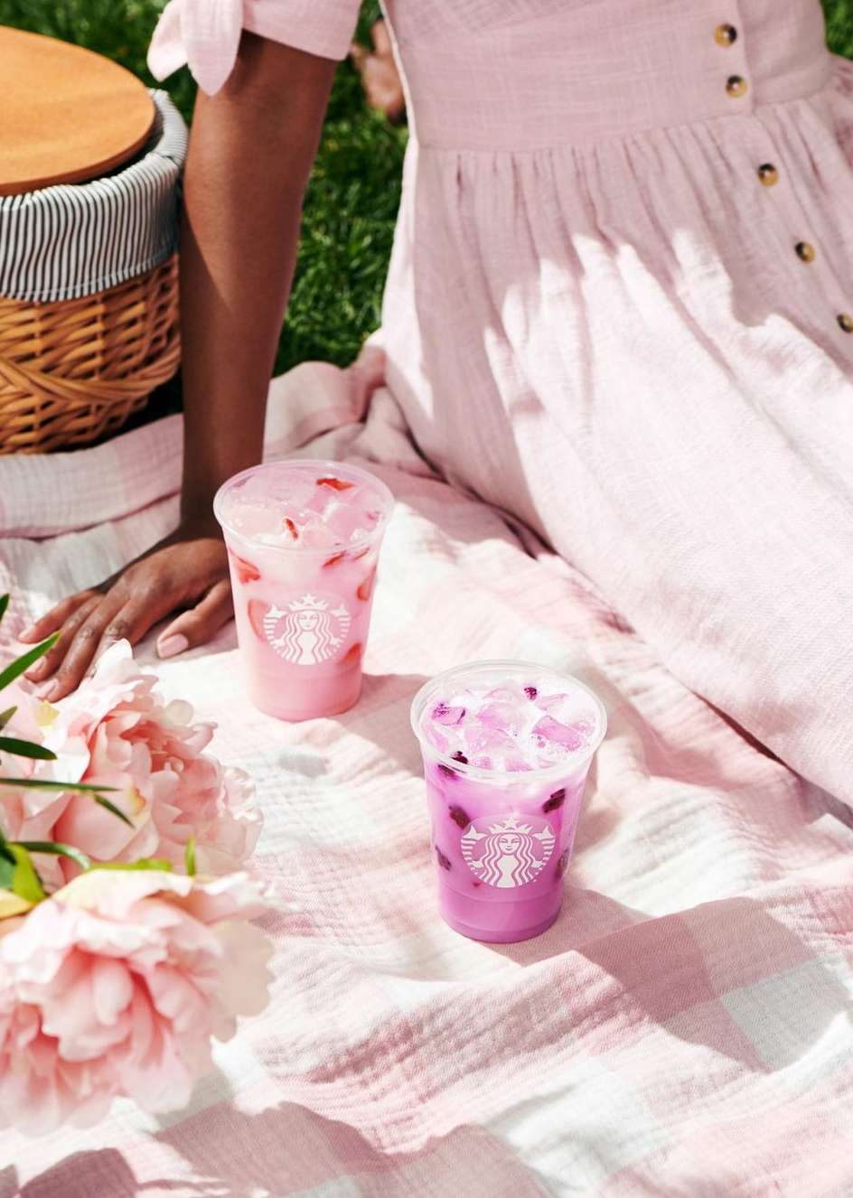 Two Starbucks Refreshers in plastic cups on a picnic blanket with pink flowers and a picnic basket on green grass outside.