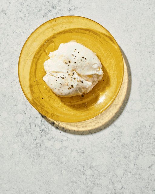 Overhead shot of burrata cheese with seasonings on a yellow plate, presented on a textured white surface in bright lighting.