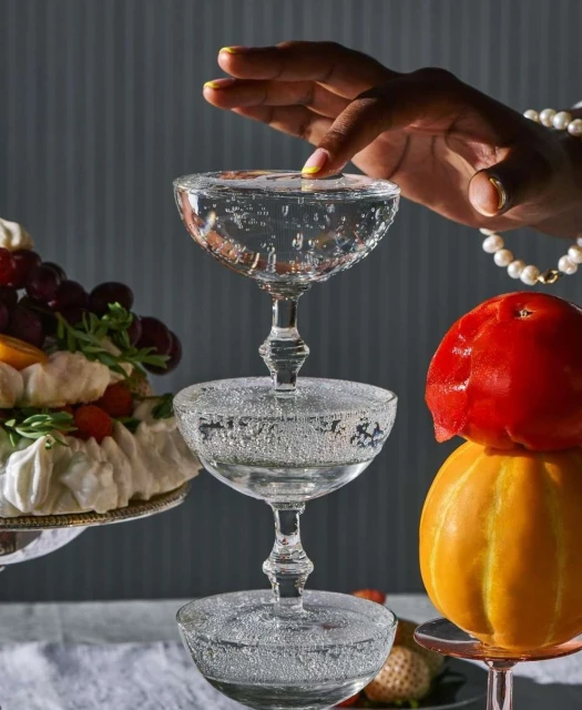 Champagne coupe tower with fruit, food still life featuring a hand touching the top glass, set against a grey background and soft lighting.