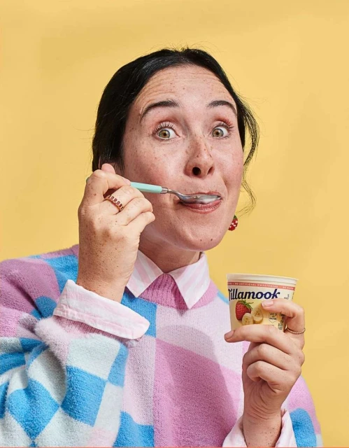 A woman smiles while holding a spoon and a container of Tillamook yogurt against a yellow background, wearing a pastel sweater.