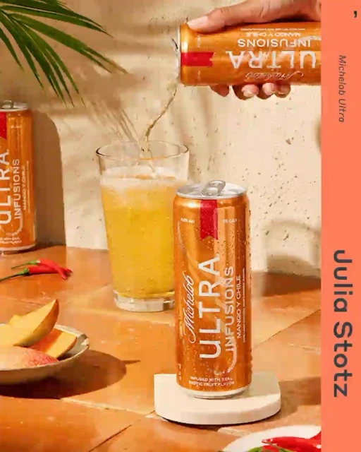 Pouring Michelob Ultra Infusions Mango y Chile beer into a glass, set against a tiled surface with mango slices and chili peppers.