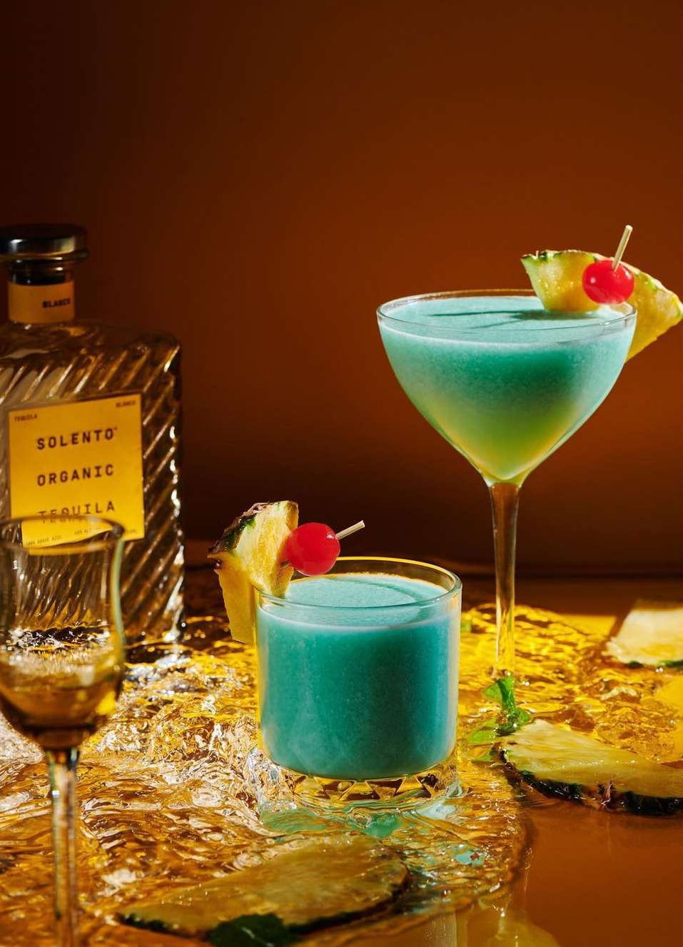 Two blue cocktails with pineapple and a cherry, beside a bottle of tequila, sitting on a reflective surface with ice.