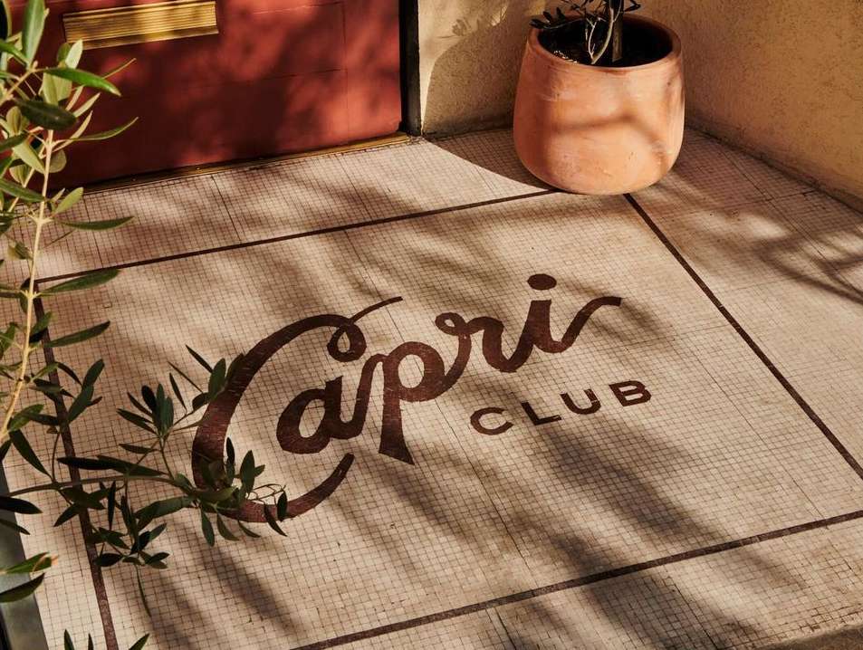 Entrance to Capri Club with terracotta tiled floor, cursive logo, and potted plant near a red door with sunlight casting shadows.