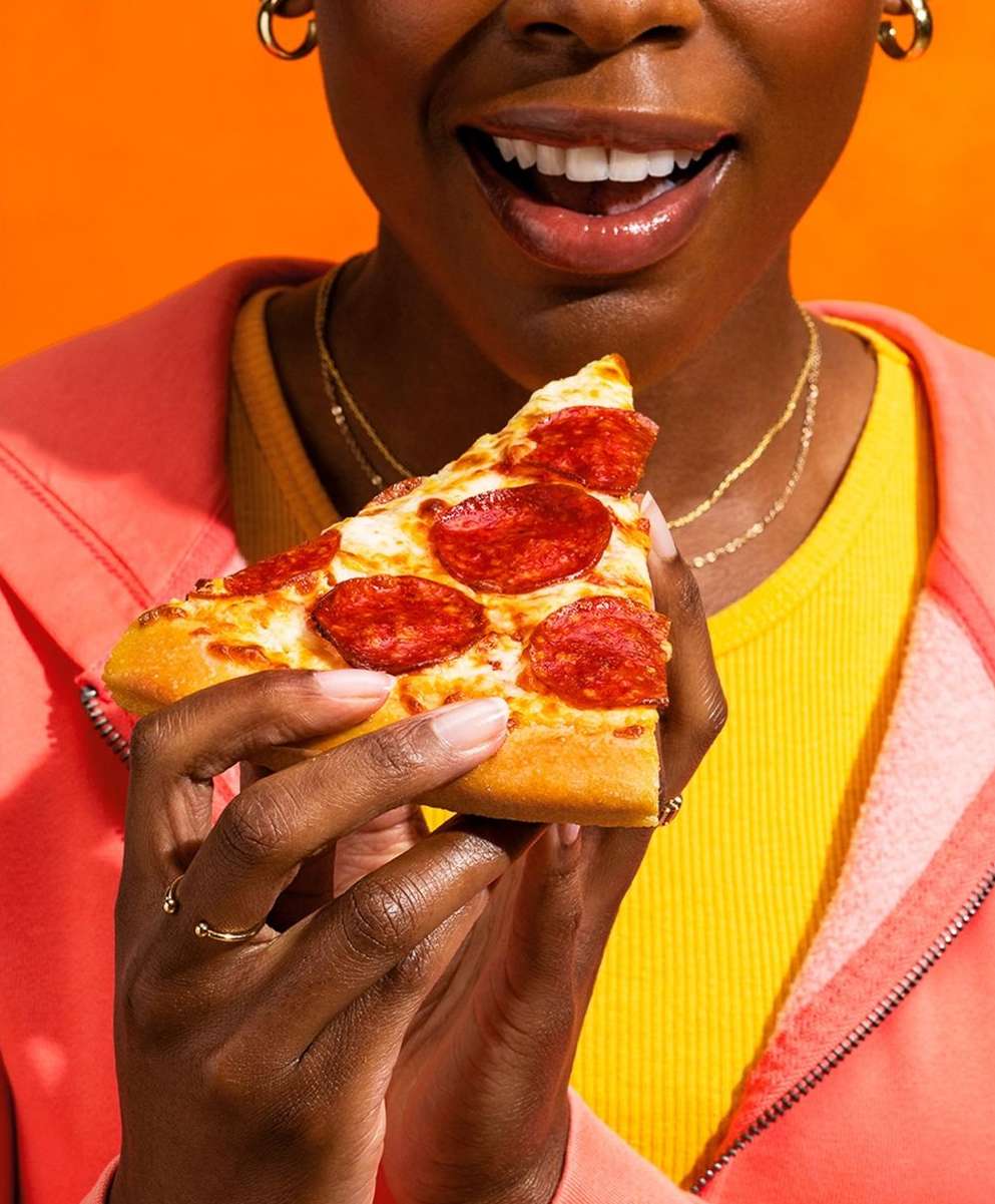 Close-up of a woman of color holding a slice of pepperoni pizza against an orange background, wearing a yellow top and pink jacket.