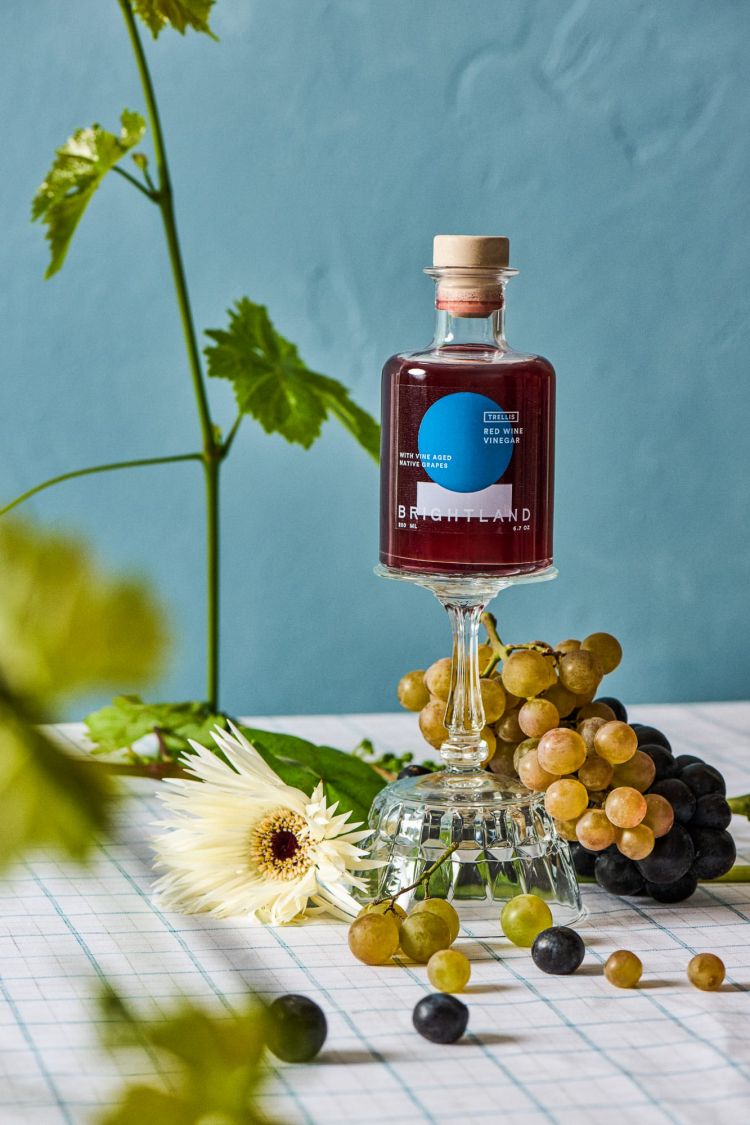 A bottle of red wine vinegar sits on a glass with grapes and a white flower against a blue background and checkered tablecloth.